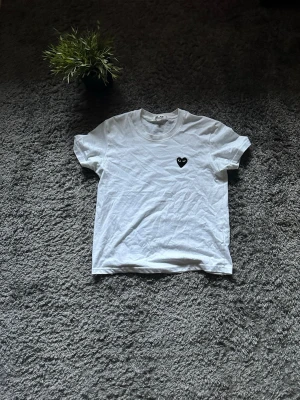 Vit Comme des Garçons t-shirt hjärta - Vit t-shirt från Comme des Garçons med klassisk svart hjärtdetalj på bröstet. T-shirten har rund hals och korta ärmar, tillverkad i mjuk bomull. Perfekt för en clean och stilren look.