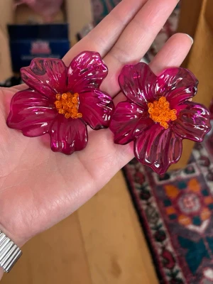 blommiga örhängen i rosa färg - Unika örhängen formade som stora blommor i transparent rosa plast med orange pärlor i mitten. Perfekta för dig som vill sticka ut och addera färg till din stil. Blommorna har en cool, lekfull vibe och är riktigt statement pieces.