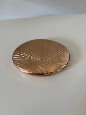 Charlotte Tilbury bronzer i guldig dosa - Snygg bronzer från Charlotte Tilbury i en lyxig, rund guldig dosa med spegel. Pudret har en varm brun nyans som ger en solkysst look. Perfekt för att framhäva ansiktskonturer och skapa glow. Dosan är elegant med räfflad yta och logga på locket.