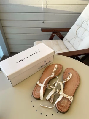 Vita sandaler från C’ M PARIS - Snygga vita sandaler från Super Mode med tunna remmar och guldfärgade detaljer. Sandalerna har dekorativa nitar och små dödskalle-berlocker på remmarna. Perfekta för sommaren och lätta att matcha till olika outfits.