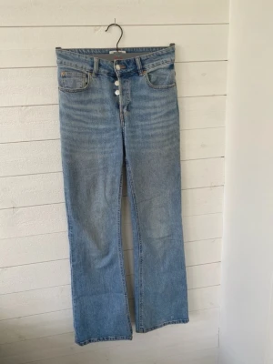 Blå raka jeans från H&M - Säljer ett par klassiska blå jeans från H&M och hög midja. Jeansen har knäppning med fyra silverfärgade knappar och femficksmodell. Materialet är slitstark bomullsdenim i en ljusblå tvätt. Perfekta till sneakers eller boots.