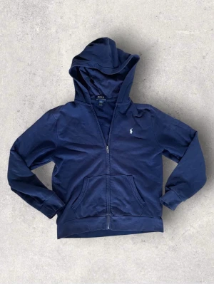 Polo Ralph Lauren mörkblå zip hoodie – stl 14–16, passar ca 158–168 cm - Mörkblå zip hoodie från Polo Ralph Lauren i storlek L/G (14–16). Klassisk modell med dragkedja, huva och två framfickor samt vit broderad logga på bröstet.  Passar ungefär någon som är ca 158–168 cm lång beroende på hur man vill att den ska sitta.  Tröjan är i mycket bra skick men har ett väldigt litet hål på ena ärmen som knappt syns när man har på sig den. Annars är den fin i färgen och fortfarande mycket fräsch. Perfekt som vardagströja eller för en stilren casual look.
