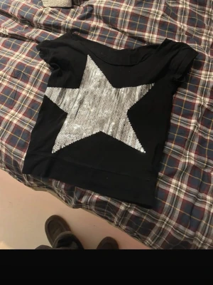 Svart topp med paljettstjärna - Snygg svart topp med en stor silverfärgad stjärna i paljetter på framsidan. T-shirten har korta ärmar och en avslappnad passform. Perfekt för dig som vill sticka ut lite extra med en glittrig detalj.