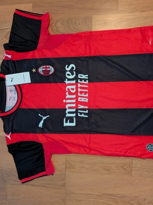 AC Milan matchtröja Puma röd/svart. Storlek. M.L - Helt ny AC Milan matchtröja från Puma i klassiska röda och svarta vertikala ränder. Tröjan har klubbmärke, Puma-logga och Emirates Fly Better-tryck på bröstet. Korta ärmar med svarta detaljer och rund hals. Perfekt för fotbollsfans!