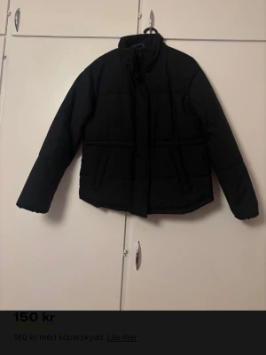 Svart pufferjacka med dragkedja - Säljer en svart pufferjacka med hög krage och dragkedja framtill. Jackan har två stora fickor på sidorna och stängs med både dragkedja och tryckknappar. Perfekt för kalla dagar och har en klassisk, stilren look.