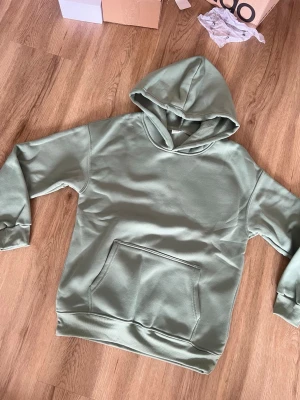 Sagegrön hoodie med magficka - En stilren sagegrön hoodie med huva och stor magficka framtill. Hoodien har ribbade muddar vid ärmslut och nederkant. Perfekt för en avslappnad look och enkel att matcha med jeans eller joggers. Materialet känns mjukt och bekvämt mot huden.