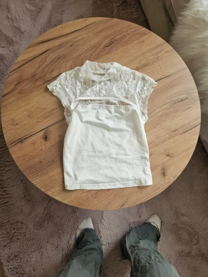Vit spetsblus med hög krage - Supersöt vit blus med spets upptill och hög krage. Ärmlös design med transparent spets över axlar och bröst, samt slät tygdel nedtill. Perfekt för en utekväll eller en fin sommardag🩷