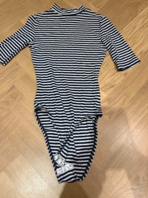 Randig body från Divided - Snygg body från Divided med marinblå och vita ränder. Modellen har halvlång ärm, hög hals och knäppning i grenen. Tillverkad i mjukt, stretchigt material som sitter skönt mot kroppen. Perfekt för en trendig och avslappnad look.