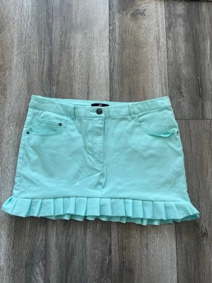 Mintgrön kortkjol med volang från H&M - Superfin mintgrön/coral blå kortkjol från H&M med volangkant nertill. Kjolen har klassiska jeansfickor fram och bak, bälteshällor och stängs med knapp och dragkedja. Perfekt för dig som vill ha en söt och fräsch look.