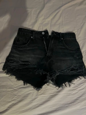 Svarta denimshorts med fransig kant - Säljer ett par svarta midwaisted jeansshorts med rå, fransig kant och slitna detaljer framtill. Klassisk femficksmodell med dragkedja och knapp. Perfekta för en avslappnad och trendig look.