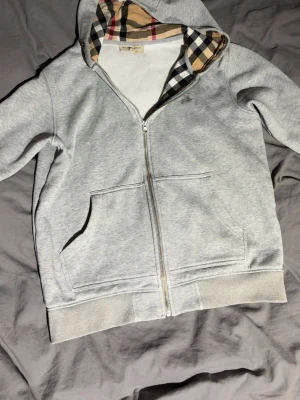 Grå hoodie Burberry hoodie - Snygg grå zip-hoodie från Burberry med klassiskt rutigt foder i huvan och insidan. Hoodien har dragkedja framtill, två fickor och ribbade muddar. Perfekt för dig som gillar stilrena plagg med en twist. Materialet är mjukt och bekvämt.