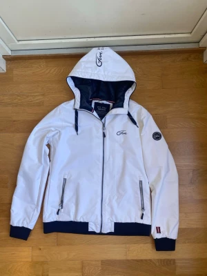 Vit vindjacka från Five Seasons - Vit vindjacka med marinblå detaljer från Five Seasons. Jackan har huva med dragsko, två fickor med dragkedja och muddar i ärmslut och nederkant. Logotyp på bröstet och ärmen. Perfekt för blåsiga dagar.