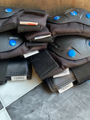 Svarta skydd från Oxelo - Säljer ett set med svarta skydd från Oxelo, perfekta för skateboard, inlines eller cykling. Setet innehåller handledsskydd, knäskydd och armbågsskydd med blå knappar och justerbara kardborreband för bästa passform. Tillverkade i slitstarkt material. Finns flera repor men i väldigt bra skick.