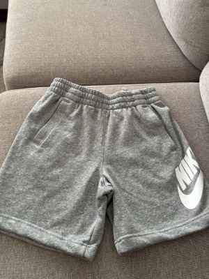 Grå mjukisshorts från Nike - Snygga grå mjukisshorts från Nike med resår i midjan och två sidofickor. Stort vitt Nike-tryck på ena benet ger en sportig vibe. Perfekta för chill eller träning, tillverkade i mjukt material för maximal komfort.