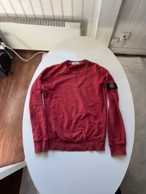 Röd långärmad tröja Stone Island - Röd långärmad tröja från Stone Island med klassisk rund halsringning och ikonisk logotyp-patch på vänster ärm. Tröjan har ribbade muddar vid ärmslut och nederkant, perfekt för en stilren och avslappnad look.