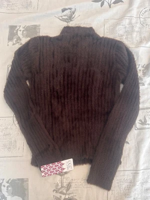 Brun fluffig stickad tröja  - Mysig brun stickad tröja  med fluffig finish och lång ärm. Tröjan har en högre krage och är tillverkad i ett mjukt material som ger en lyxig känsla. Perfekt för dig som gillar en trendig och bekväm look.