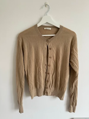 Beige Malene Birger kofta med rund hals - Klassisk beige kofta med rund halsringning och knappar framtill. Långärmad modell med ribbade muddar vid ärmslut och nederkant. Tillverkad i ett tunt, mjukt material.