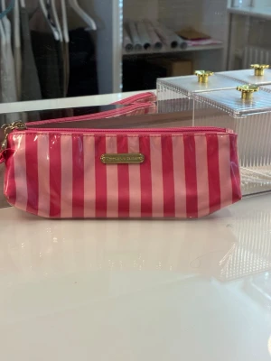 Rosa randig necessär Victoria's Secret - Säljer en snygg necessär från Victoria's Secret med rosa och ljusrosa vertikala ränder. Väskan har en blank yta i plastmaterial, dragkedja upptill och en liten metallskylt med loggan på framsidan. Perfekt för smink eller småprylar. Säljs även som pennfack, så den kan användas även för detta ändamål.😊Den är i perfekt skick!