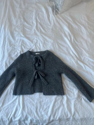 Mörkgrå stickad cardigan med knyt - Snygg mörkgrå stickad cardigan med långa ärmar och trendig knytning framtill. Kortare modell som passar perfekt till högmidjade jeans eller kjol. Mjuk och skön kvalitet, perfekt för lager på lager under kyliga dagar. Oanvänd. Nypris: 499kr