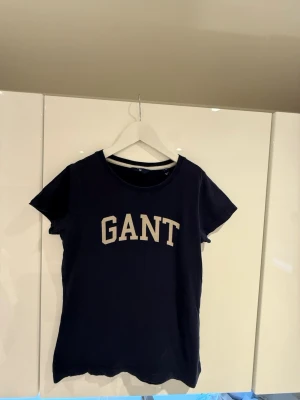 Marinblå GANT t-shirt med tryck - Snygg marinblå t-shirt från GANT med klassiskt vitt tryck framtill. T-shirten har rund halsringning och korta ärmar. Tillverkad i mjuk bomull som känns skön mot huden. Perfekt för en avslappnad och stilren look.