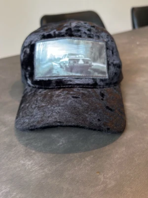 Svart meshkeps med bilmotiv - Unik svart keps i sammetsliknande material framtill och mesh baktill. Framtill finns en stor patch med ett coolt bilmotiv i svartvitt. Perfekt för dig som vill sticka ut med en annorlunda accessoar.