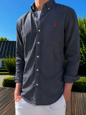 Mörkblå/grå skjorta från Ralph Lauren - Klassisk mörkgrå skjorta från Ralph Lauren med knappslå och vit knappstängning. Skjortan har lång ärm och den ikoniska röda logotypen broderad på bröstet. Tillverkad i bomull och har en normal passform. Perfekt för en clean och stilren look.