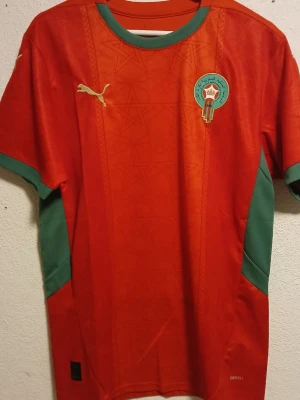 Marocko fotbollströja Puma röd/grön - Snygg röd fotbollströja från Puma med Marockos landslagsemblem på bröstet och guldfärgad Puma-logga. Gröna detaljer vid ärmar och sidor samt rund halsringning. Tillverkad i lätt och ventilerande material, perfekt för träning eller match. I nytt skick
