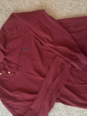 Vinröd långärmad henley från Polo Ralph Lauren - Säljer en vinröd långärmad henley från Polo Ralph Lauren med klassisk blå logga broderad på bröstet. Skjortan har två knappar vid halsen och är gjord i mjuk bomull. Perfekt för en avslappnad och stilren look.