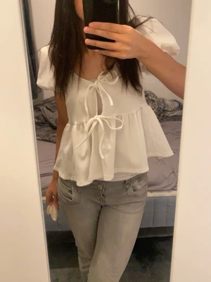 Vit topp med puffärm och knytband - Supersöt vit topp med puffärmar och två knytband framtill. Modellen är luftig och har en lätt volang nedtill som ger en romantisk vibe. Perfekt till jeans eller kjol. Materialet ser ut att vara tunt och flowy, troligen bomull eller viskos.