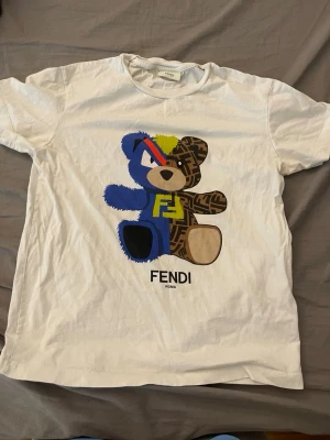 Vit Fendi t-shirt med björnmotiv (Kids)  - Vit t-shirt från Fendi (Kids, 12+) med ett stort färgglatt björnmotiv på framsidan. Björnen har blå, brun och gula detaljer samt Fendi-logga. T-shirten har rund hals och korta ärmar, perfekt för en trendig och lekfull stil.