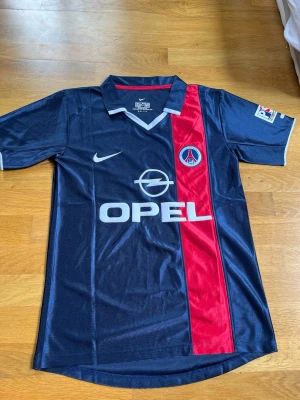 PSG fotbollströja Ronaldinho #21 - Säljer Retro 2001/2002 Mörkblå PSG matchtröja från Nike med röd vertikal rand, OPEL sponsortryck och klubbmärke på bröstet. På ryggen står det 'RONALDINHO' och nummer 21 i rött och vitt. Kortärmad med krage, tillverkad i polyester.