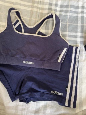 Mörkblått träningsset från Adidas - Snyggt mörkblått träningsset från Adidas med sport-bh och shorts. Setet har vita detaljer och klassiska Adidas-ränder på shortsen. Perfekt för gymmet eller löpning. Materialet är mjukt och stretchigt för maximal komfort.