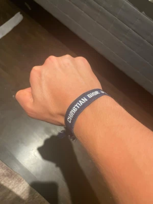 Blått Christian Dior armband - Snyggt blått armband från Christian Dior med vit broderad text. Armbandet är gjort i tyg och har fransade kanter för en avslappnad vibe. Perfekt accessoar för dig som vill addera lite lyx till din outfit.
