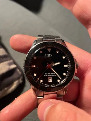 Tissot Seastar klocka i silver - Säljer en Tissot Seastar klocka med svart urtavla, datumvisning och röd sekundvisare. Klockan har en robust boett och länk i silverfärgat stål. Vattentät upp till 300 meter och har tydliga indexmarkeringar. Perfekt för dig som gillar stilrena accessoarer.