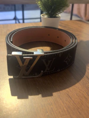 Bälte - Snyggt svart bälte med klassiskt monogrammönster i silver på spännet. Skärpet är gjort i skinn och har en exklusiv känsla som lyfter vilken outfit som helst.
