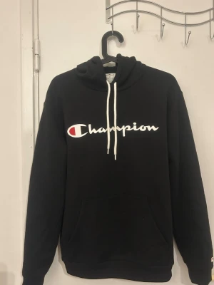 Svart Champion hoodie med logga - Säljer en svart hoodie från Champion med stor vit logga och röd detalj på bröstet. Hoodien har huva med vita snören och en stor magficka. Mjuk bomullskänsla och ribbade muddar vid ärmar och nederkant. Perfekt för en avslappnad och sportig stil.