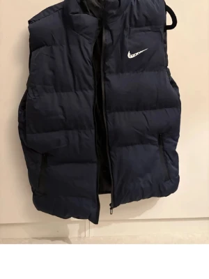 Mörkblå dunväst från Nike - Snygg mörkblå dunväst från Nike med dragkedja framtill och två sidofickor med dragkedja. Västen har en hög krage och den klassiska Nike-loggan på bröstet. Perfekt för lager-på-lager och streetwear-stil.