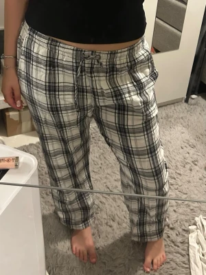 Rutiga pyjamasbyxor i vitt och svart - Säljer ett par mysiga pyjamasbyxor med svartvitt rutmönster och dragsko i midjan. Byxorna har raka ben och är perfekta för chill dagar hemma. Tillverkade i mjukt tyg som känns skönt mot huden.