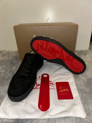 Svarta sneakers från Christian Louboutin - Stilrena svarta sneakers i mocka från Christian Louboutin med den ikoniska röda sulan och diskret logga på sidan. Skorna har svarta skosnören och en klassisk låg siluett. Perfekta för dig som vill ha en exklusiv touch på din outfit.