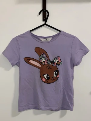 Lila t-shirt med kanintryck - Söt lila t-shirt med ett stort tryck av en brun kanin med blommig rosett framtill. T-shirten har rund halsringning och korta ärmar. Perfekt för dig som gillar gulliga motiv och vill ha en färgglad touch i garderoben.