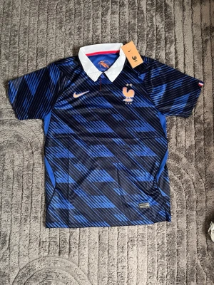 Frankrike fotbollströja Nike blå - Snygg blå fotbollströja från Nike med Frankrikes landslagsmärke på bröstet. Tröjan har ett mönster i olika blå nyanser, vit krage och orange Nike-logga. Tillverkad i lätt och ventilerande material, perfekt för träning eller match.