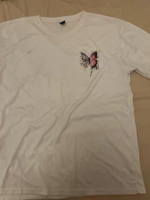Vit t-shirt med rosa fjärilstryck - Vit t-shirt med ett stort tryck av en rosa och svart fjäril på ryggen och ett mindre likadant tryck på bröstet. Klassisk rund halsringning och korta ärmar. Perfekt för dig som gillar unika och konstnärliga motiv.