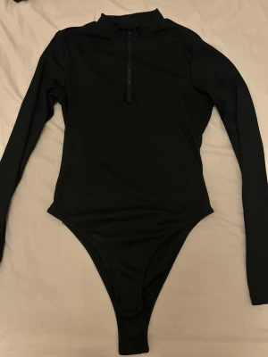 Svart ribbad body med dragkedja - Snygg svart body i ribbat material med lång ärm och hög krage. Dragkedja framtill som går ner till bröstet för en sportig look. Perfekt att styla med jeans eller kjol för en trendig outfit.