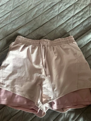 Ljusrosa träningsshorts med snörning - Snygga ljusrosa träningsshorts med elastisk midja och snörning framtill. Shortsen har dubbla lager för extra komfort och en sportig look. Perfekta för gymmet eller löprundan.
