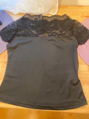 Svart topp med tull helt ny - Snygg svart topp med korta ärmar och vacker spets upptill över axlar och bröst. Toppen har en feminin och elegant look med rund halsringning och mjukt, stretchigt material.  Jag vill säga eftersom den var för liten. Jag fick den som present från min familj. Helt ny. 