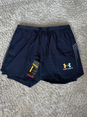 Svarta träningsshorts Under Armour - Snygga svarta träningsshorts från Under Armour med färgglada prickdetaljer på sidorna och logga i regnbågsfärger. Shortsen har snörning i midjan och är tillverkade i ett lätt och snabbtorkande syntetmaterial. Perfekta för gymmet eller löprundan.