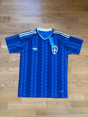 Blå Sverige fotbollströja Adidas - Säljer en blå fotbollströja från Adidas med Sveriges landslagsemblem på bröstet. Tröjan har ett unikt mönster i ljusblått, vita ränder på axlarna och vit krage med gul detalj. Perfekt för match eller träning.