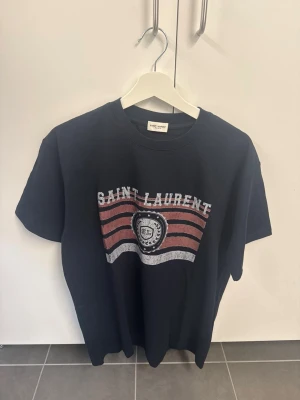 Mörkblå Saint Laurent t-shirt med tryck - Cool mörkblå t-shirt från Saint Laurent med stort vintageinspirerat tryck i vitt och rött framtill. Klassisk rund halsringning och korta ärmar. Tillverkad i mjuk bomull som känns skön mot huden. Perfekt för dig som vill ha en statement tee med lyxig känsla.