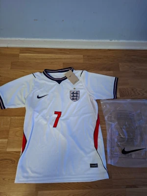 England fotbollströja Nike Saka #7 VM 26/27 - England landslagets vita fotbollströja från Nike med Saka och nummer 7 på ryggen. Tröjan har svarta och röda detaljer, broderad landslagssköld och Nike-logga på bröstet. Tillverkad i lätt och ventilerande material, perfekt för match eller träning.