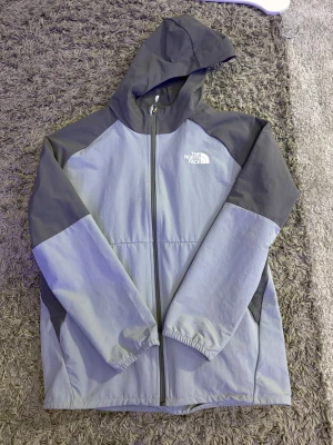 Grå vindjacka från The North Face - Snygg tvåfärgad vindjacka från The North Face i ljusgrått och mörkgrått. Jackan har huva, dragkedja framtill och elastiska muddar vid ärmsluten. Tillverkad i lätt och tåligt material, perfekt för blåsiga dagar. Logga på bröstet och ryggen.